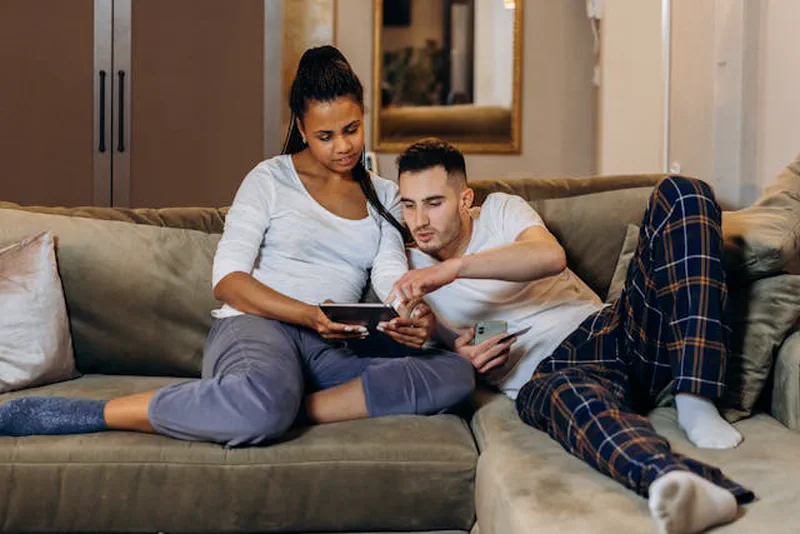 Couple sitting together on a sofa looking at a tablet and phone — planning a meaningful evening at home